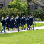 Pending graduates wearing dark blue gowns walk on a sunny spring day Penn State's University Park campus.