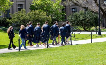 Pending graduates wearing dark blue gowns walk on a sunny spring day Penn State's University Park campus.