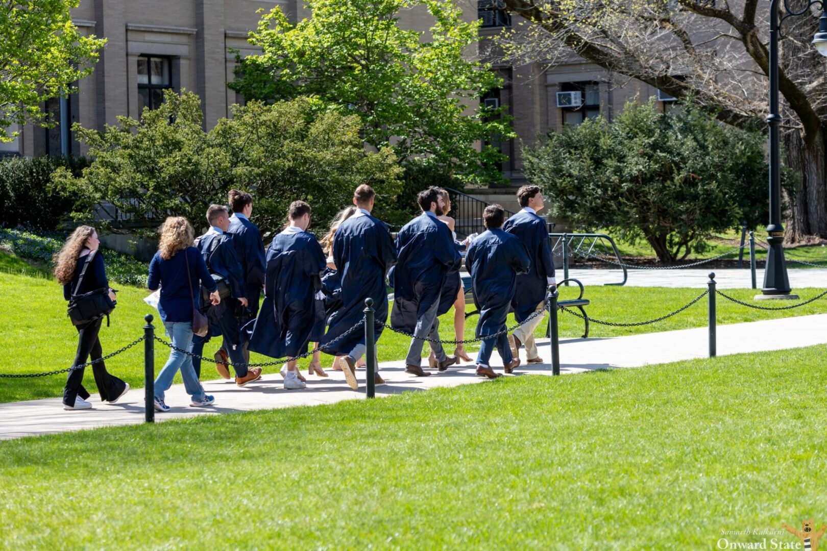 Pending graduates wearing dark blue gowns walk on a sunny spring day Penn State's University Park campus.