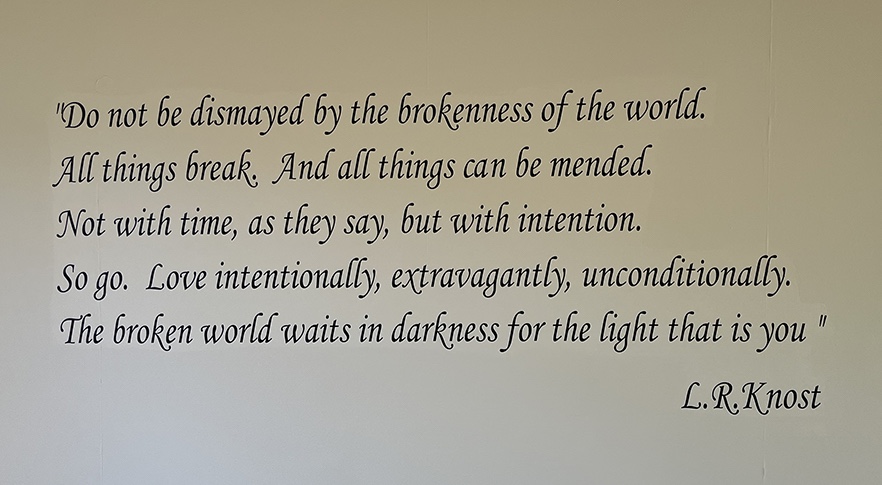 A sign on a wall shows the L.R. Knost quote "“Do not be dismayed by the brokenness of the world. All things break. And all things can be mended. Not with time, as they say, but with intention. So go. Love intentionally, extravagantly, unconditionally. The broken world waits in darkness for the light that is you.”