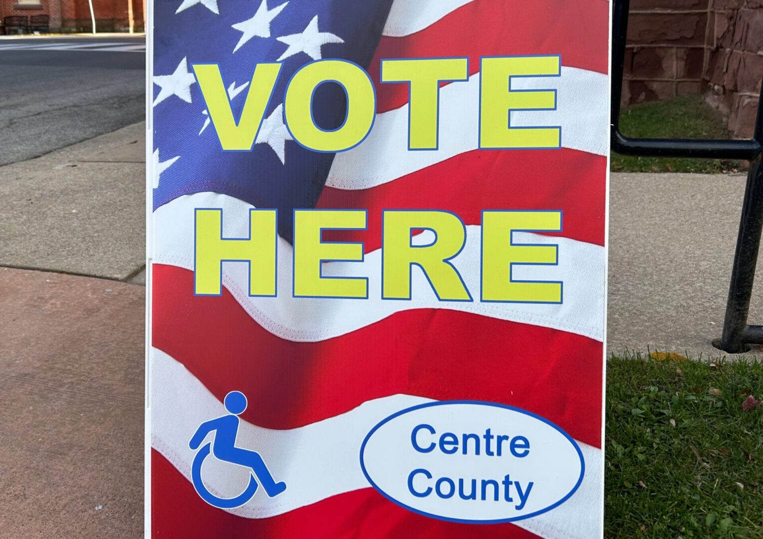 VOTE HERE in yellow letters, Centre County and a disability symbol against an American flag background on a sandwich board in Bellefonte, PA.