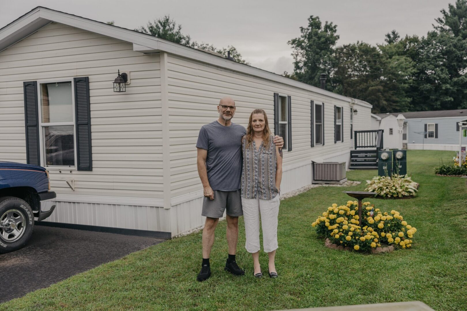 A man and woman stand in front of a mobile home with yellow flowers next to them.