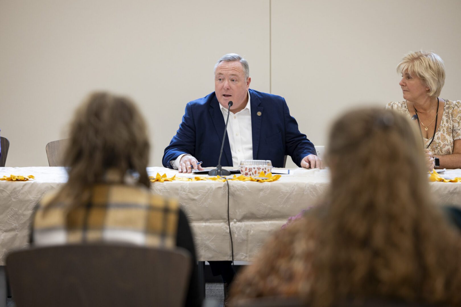 Aging Secretary Jason Kavulich speaks into a microphone during a meeting, with attendees in the foreground and a woman seated beside him.